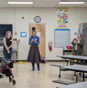 Three caucasian appearing women stand left to right in a school cafeteria cafeteria. The woman in the center has a microphone and is speaking to an audience that is not visible.