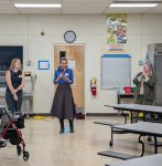 Three caucasian appearing women stand left to right in a school cafeteria cafeteria. The woman in the center has a microphone and is speaking to an audience that is not visible.